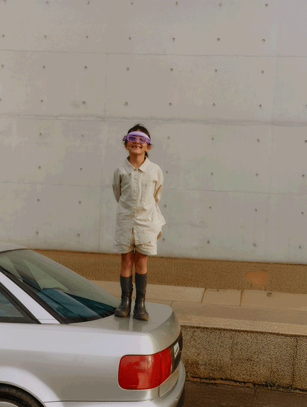 Child in boots, standing on the back of a car against a concrete wall.