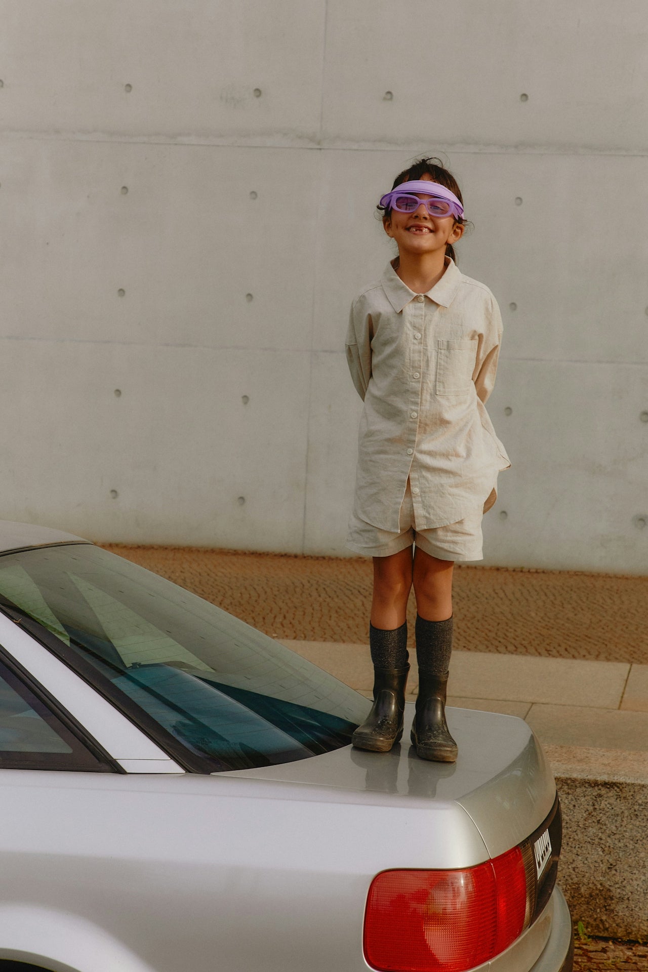 Child in boots, standing on the back of a car against a concrete wall.