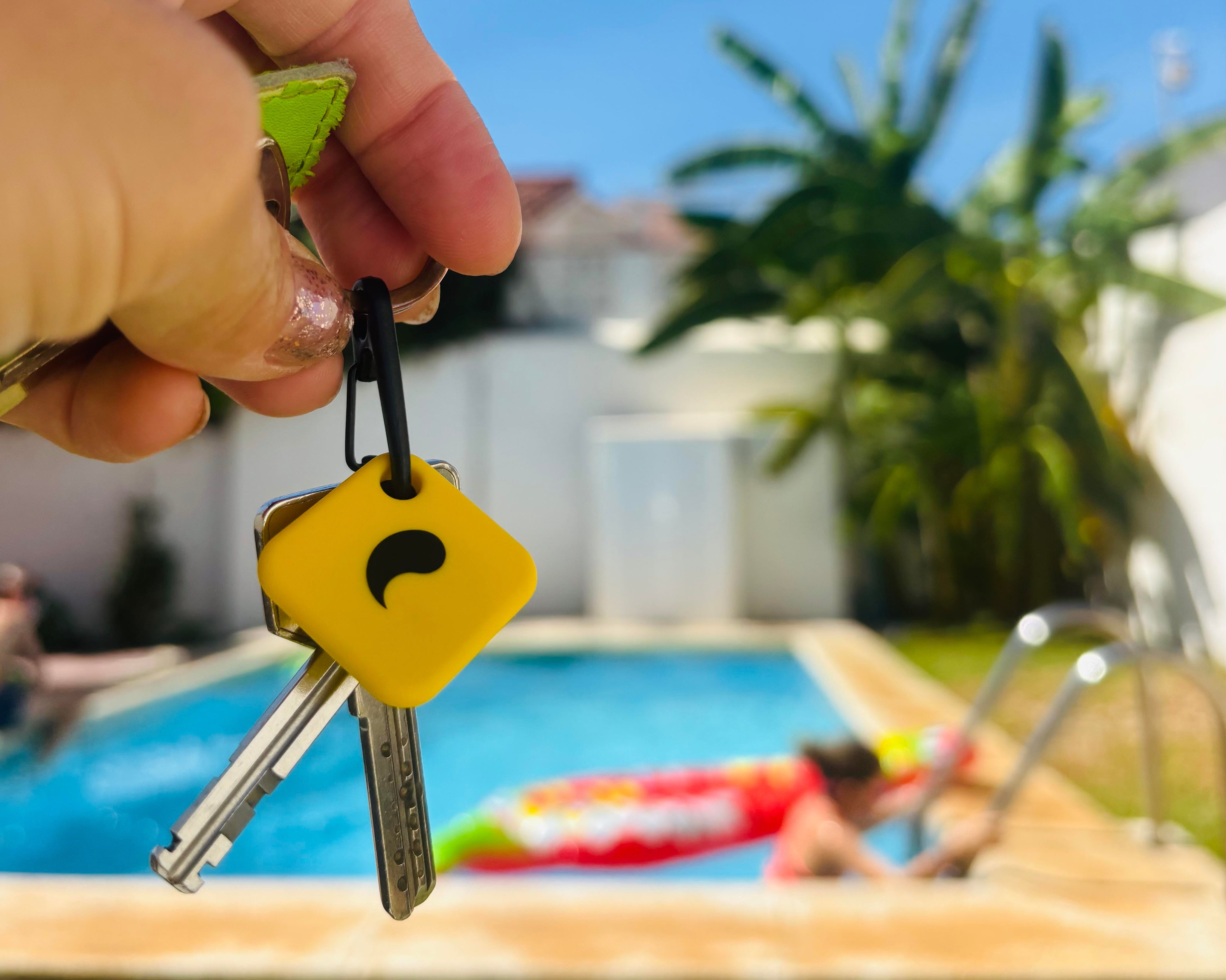 A hand holding a set of keys with the unpluq tag in the foreground, with a blurred outdoor poolside background under a blue sky.